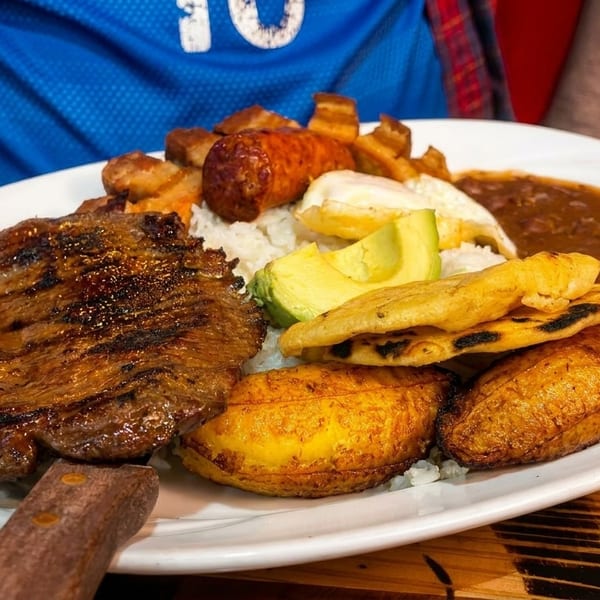 A large platter of grilled steak, rice, beans, avocado, plantains, sausage, and fried egg.