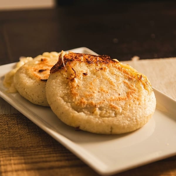 Two golden-brown arepas with melted cheese on a white rectangular plate, on a woven placemat.
