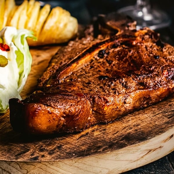 A grilled T-bone steak, wedge salad with dressing, and a baked potato on a wooden board.