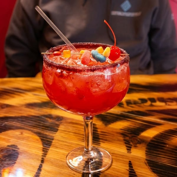 Red cocktail in a goblet, garnished with colorful candies and a cherry, on a wooden table.