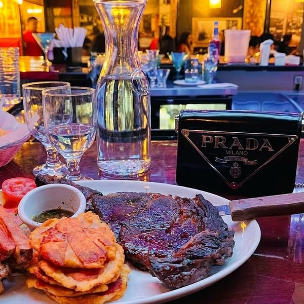 Grilled steak with fries, plantains, and a mojito on a bar counter with a Prada bag.